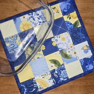 Könnte beinhalten: Ein quadratischer, gesteppter Topflappen mit einem Patchwork-Design aus blauen und gelben Stoffquadraten mit Blumen-, Paisley- und Unimustern sowie einem dunkelblauen Rand, teilweise bedeckt von einer klaren Glasauflaufform mit geschwungenen Rändern.