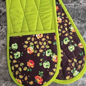 May include: A double oven glove featuring a quilted lime green upper section and a dark brown lower section decorated with a floral and paisley pattern in shades of orange, yellow, and green. The entire piece is finished with lime green trim.