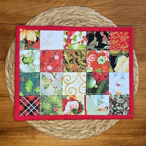May include: A rectangular quilted fabric mat featuring a patchwork design of twenty squares with various Christmas-themed patterns, including holly, poinsettias, candy canes, a snowman, and Santa Claus, all bordered by a solid red fabric binding.