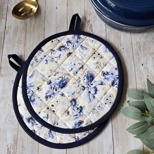 May include: Two round quilted pot holders featuring a cream-coloured fabric with a blue floral pattern. Each pot holder has a dark navy blue fabric trim around the edge and a small fabric loop at the top for hanging, with a diamond-stitched texture.