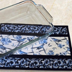 May include: A clear glass baking dish sits on a quilted placemat. The placemat has a blue and white bird and floral design with a black border. The design includes blue birds perched on branches with blue flowers.