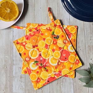 Peut inclure: Un dessous de plat carré matelassé orné d'un motif d'agrumes coloré avec des tranches d'oranges, de pamplemousses et de citrons dans des tons jaune, orange et rose, agrémenté de petites baies rouges et de feuilles vertes, avec une boucle de suspension.