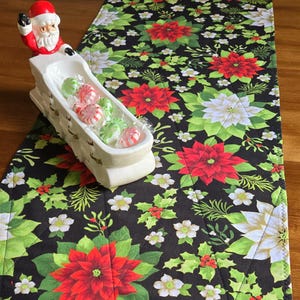 May include: A festive table runner with a black backdrop, adorned with red and white poinsettias, green foliage, and white blossoms. A white ceramic Santa Claus sleigh, filled with wrapped sweets, is placed on the runner.
