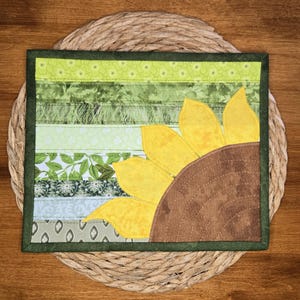 Peut inclure: Un quilt rectangulaire en tissu présentant un fond à rayures horizontales composé de divers tissus verts à motifs. Un grand motif de tournesol partiel avec des pétales jaune vif et un centre brun texturé est appliqué sur le côté droit.