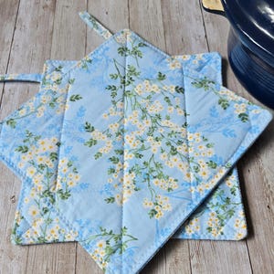 May include: Two square quilted pot holders featuring a light blue fabric base with a repeating pattern of small white flowers and green leaves. Each pot holder has a fabric loop attached to one corner for hanging and visible stitched seams.