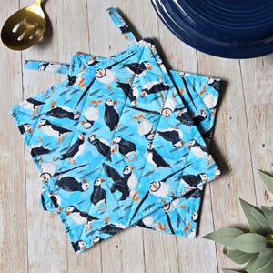 May include: A square quilted potholder featuring a repeating pattern of black and white puffins with orange beaks and feet on a bright blue background. The fabric has a stitched diamond grid pattern and includes a small fabric loop for hanging.