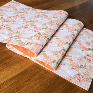 May include: A quilted table runner featuring a white fabric top with a repeating pattern of orange and peach-coloured hydrangea flowers and green leaves. The underside of the runner is a solid orange fabric with a textured, dimpled surface finish.
