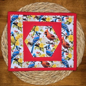 May include: A square quilted potholder featuring a central hexagon panel surrounded by a red fabric border. The fabric displays a pattern of blue jays and red cardinals perched among yellow flowers and green leaves on a white background.