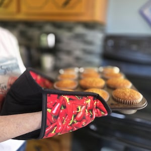 May include: An oven glove featuring a vibrant red chilli pepper pattern on a black background, with black trim along the edges. The glove is shown holding a metal muffin tin containing several golden-brown baked muffins in paper liners.