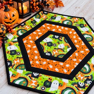May include: A hexagonal quilted table mat featuring a repeating Halloween pattern of white ghosts, green spiders, and black skulls on bright orange and lime green backgrounds, separated by solid black fabric borders and finished with black binding.