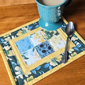May include: A rectangular quilted placemat featuring a central grid of six fabric squares in shades of blue, yellow, and white, surrounded by a dark green floral border and a solid yellow trim. A teal mug and a silver spoon rest on the placemat.