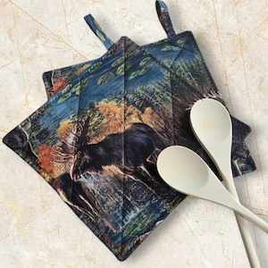 May include: Two square quilted pot holders with a forest scene featuring a moose. The pot holders have a dark blue border and a loop for hanging. Two wooden spoons are next to the pot holders.