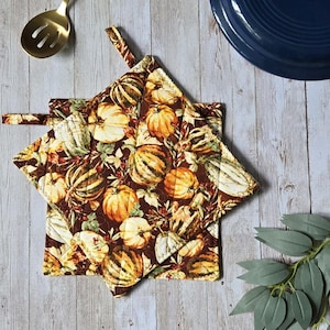 May include: Two square quilted pot holders featuring a dark brown background with a repeating pattern of orange, yellow, and green pumpkins, gourds, and autumn leaves. Each pot holder includes a small fabric loop for hanging in the kitchen.