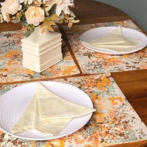 Peut inclure: Deux sets de table rectangulaires avec un motif floral dans des tons orange, marron et bleu sarcelle sur fond crème. Chaque set porte une assiette blanche avec une serviette crème pliée. Un vase blanc avec des fleurs pêche est posé.