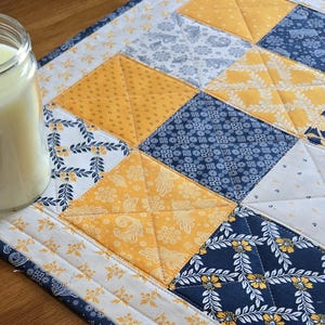 May include: A quilted table runner with a patchwork design featuring yellow and blue fabric squares with various patterns. The runner has a white border and is made with a variety of fabrics.