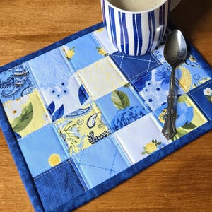 May include: A rectangular quilted placemat featuring a patchwork design of blue, white, and yellow fabric squares with floral, paisley, and geometric patterns. A white mug with blue vertical stripes and a silver metal spoon rest on the placemat.