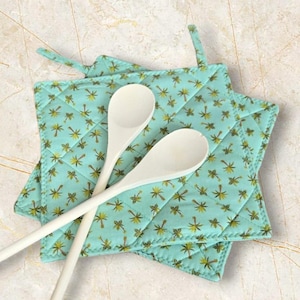 May include: Two turquoise blue quilted pot holders with a palm tree pattern and two wooden spoons on top.