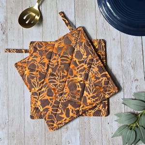 May include: A square quilted pot holder featuring a brown and orange batik-style leaf pattern. The fabric has a diamond-stitched texture and includes a small fabric loop at the corner for hanging. The design displays a repeating organic motif.