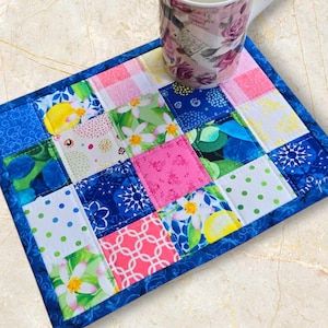 May include: A patchwork coaster with a blue border, featuring squares of various patterns including floral, polka dots, and geometric designs. A floral mug rests on the coaster. The coaster is approximately 15 cm square.