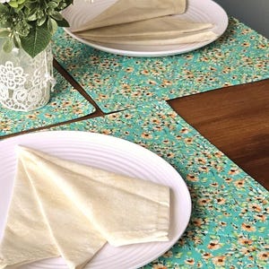 Peut inclure: Un ensemble de sets de table rectangulaires présentant un fond sarcelle avec un motif dense de petites fleurs orange et blanches. Chaque set de table porte une assiette blanche à stries circulaires et une serviette en tissu crème pliée.