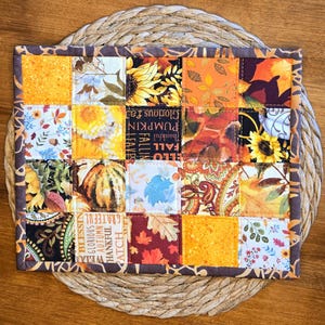 Könnte beinhalten: Ein rechteckiger Topflappen aus Stoff mit einem Patchwork-Design aus zwanzig Quadraten mit herbstlichen Mustern, darunter Sonnenblumen, Kürbisse, Herbstblätter und der Text "Grateful", "Thankful" und "Glorious Autumn" in warmen Farben.