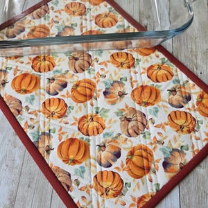 Peut inclure: Un dessous de plat rectangulaire en tissu matelassé avec un motif répétitif de citrouilles orange et de feuilles vertes sur fond blanc, bordé d'un tissu rouge foncé, placé sous un plat de cuisson rectangulaire en verre transparent.