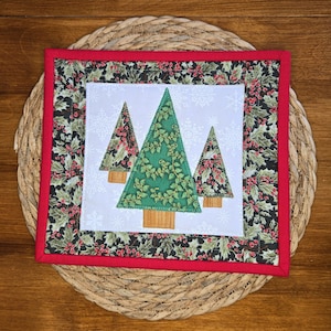 May include: A square quilted potholder featuring a central white panel with three appliqued Christmas trees in green and patterned fabric. The border is a dark fabric with a holly and berry print, finished with a bright red fabric binding.