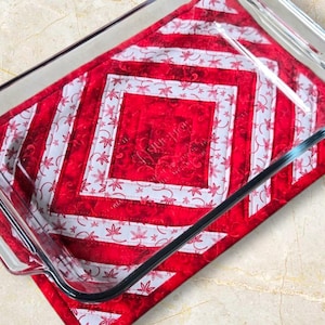 Könnte beinhalten: Eine transparente Glasbackform liegt auf einem rot-weiß gesteppten Topflappen. Der Topflappen hat ein geometrisches Design mit roten und weißen Streifen und floralen Mustern. Die Form ist leer und bereit.