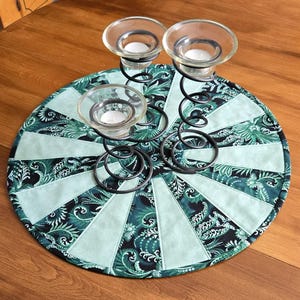 Peut inclure: Un bougeoir décoratif avec trois coupelles en verre sur une structure en métal noir. Le bougeoir est posé sur un dessous-de-plat rond et matelassé avec un motif floral vert d'eau et vert foncé. Le dessous-de-plat mesure environ 30 cm de diamètre.