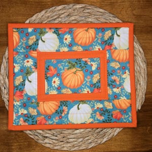 May include: A rectangular fabric potholder featuring a teal background with a repeating pattern of orange pumpkins, white pumpkins, and autumn leaves. The piece is framed by a bright orange fabric border and includes a central rectangular panel.