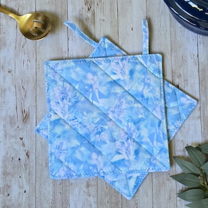 May include: Two square quilted pot holders featuring a light blue and white abstract geometric pattern. Each pot holder has a fabric loop attached to one corner for hanging. The fabric is stitched with a diamond pattern across the surface.