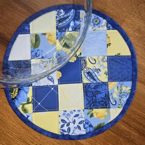 Könnte beinhalten: Ein runder, gesteppter Stoffuntersetzer mit einem Patchwork-Design aus blauen und gelben Blumen-, Paisley- und geometrischen Mustern. Die kreisförmige Matte ist mit einer dunkelblauen Stoffeinfassung versehen und wird teilweise von einer Glasschüssel bedeckt.