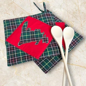 May include: Two green and red plaid pot holders with a red fabric cutout of the state of Massachusetts. The pot holders are on a white surface with two wooden spoons.