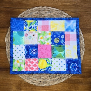 May include: A rectangular quilted fabric mat featuring a patchwork design of sixteen squares with various patterns including lemons, pink gingham, blue floral motifs, green leaves, and polka dots, all framed by a solid dark blue fabric border.
