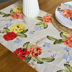 May include: Cream-coloured quilted table runner with a floral pattern featuring red, yellow, and orange roses, green leaves, and small blue flowers. The runner is rectangular and has a diamond-stitched pattern.
