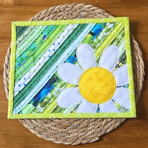 May include: A rectangular quilted fabric mat featuring diagonal strips of green, white, and blue patterned fabric. A large white daisy with a bright yellow centre is appliqued onto the lower right corner, framed by a light green border.