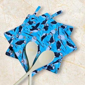 May include: A blue, star-shaped potholder patterned with puffins. Two wooden spoons are placed on the potholder. The puffins are black, white, and orange, set against a light blue background. The potholder has a hanging loop.