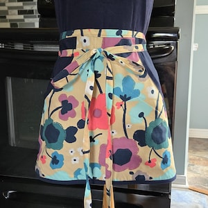 Könnte beinhalten: Eine Halbschürze mit beigem Hintergrund und einem großflächigen Blumenmuster in Marineblau, Hellblau, Koralle und Violett. Die Schürze hat einen einfarbigen marineblauen Bund und einen passenden Saum am unteren Rand sowie ein Bindeband.