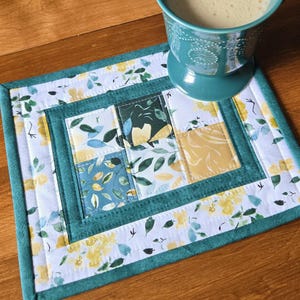 May include: A square, quilted placemat with a floral design in shades of yellow, blue, and green. The placemat has a teal border and a central patchwork pattern. A teal mug with a beverage sits on the placemat.