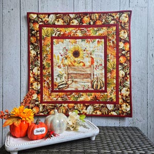 May include: A square quilted wall hanging featuring a central panel of a sunflower and pumpkins in a wooden crate, surrounded by a border of autumn leaves and pumpkins. Below, a white tray holds a small orange pumpkin and a white pumpkin.