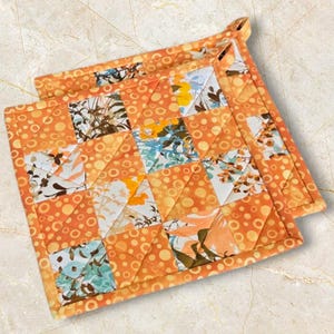 Könnte beinhalten: Zwei quadratische, gesteppte Topflappen. Sie haben ein Patchwork-Design mit orangefarbenen, weißen und blauen Blumenmustern. Der Rand ist orange mit einem gepunkteten Muster. Eine Schlaufe ist zum Aufhängen angebracht.