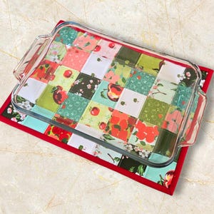 May include: A red and green patchwork hot pad with a glass baking dish on top. The hot pad is made of fabric squares with a variety of apple-themed prints.