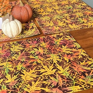 Peut inclure: Deux sets de table rectangulaires en tissu arborant un motif répétitif de feuilles d'automne dans des tons jaune, orange et vert sur un fond marron foncé, accompagnés d'une citrouille marron texturée et d'une figurine de citrouille blanche.