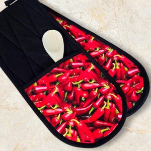 May include: A pair of black oven mitts with a red chilli pepper print. The mitts have a pocket on the side to hold a wooden spoon.