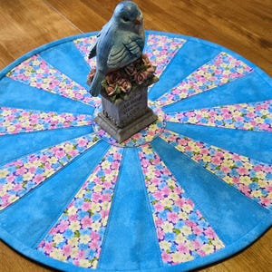 Peut inclure: Un dessous de plat rond et bleu matelassé avec un motif floral et une figurine d'oiseau bleu. Le dessous de plat présente un motif en forme d'éventail avec des sections alternées de tissu bleu uni et floral. La figurine d'oiseau est posée sur un petit socle avec l'inscription "All things grow with LOVE".