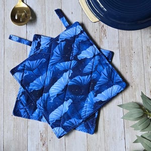 May include: Two square quilted pot holders featuring a dark blue fabric with a repeating pattern of light blue ginkgo leaves and small white dots. Each pot holder includes a small fabric loop at the corner for hanging, showcasing a stitched design.
