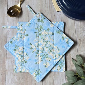May include: Two square quilted pot holders featuring a light blue background with a repeating pattern of small white flowers and green leaves. Each pot holder includes a fabric loop for hanging, with visible stitching forming a diamond grid pattern.