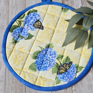 May include: A round quilted potholder featuring a pale yellow background with a subtle pattern, decorated with clusters of blue hydrangea flowers and black and yellow butterflies. The circular fabric piece is finished with a solid blue border.