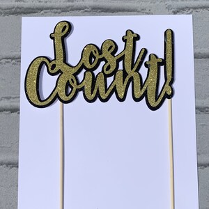 May include: Gold glitter cake topper with black outline that reads "Lost Count!" on two wooden sticks. The topper is on a white background with the text "watermelon custom designed accessories" at the bottom.