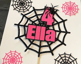 May include: A pink and black spiderweb cake topper with a pink number 4, the name "Ella" and a black spider. The topper is on a wooden stick and is surrounded by smaller spiderwebs.